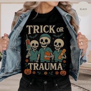 May include: Black t-shirt with the words "TRICK OR TRAUMA" and a graphic of three skeleton doctors. One skeleton holds a syringe, another a clipboard, and the third a bandage. Bats, pumpkins, and flowers complete the Halloween design.