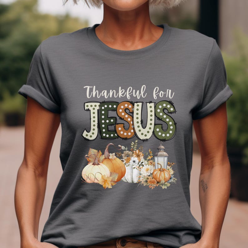 Op de afbeelding: Een donkergrijs T-shirt met de tekst "Thankful for JESUS" in een kleurrijk, gestippeld lettertype. De tekst wordt omringd door herfstthema-beelden, waaronder pompoenen, bladeren en een lantaarn.