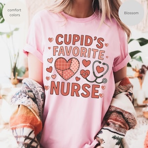 Peut inclure: T-shirt rose clair avec l'inscription "Cupid's Favorite Nurse" en rouge. Le motif comprend un cœur en patchwork, des petits cœurs et un stéthoscope. Les manches sont retroussées.