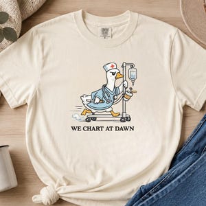 May include: A cream-colored short-sleeve t-shirt featuring a graphic of a white duck dressed as a nurse, wearing a cap with a red cross and a stethoscope, riding an IV pole on wheels while holding a coffee cup. The text reads WE CHART AT DAWN.