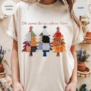 May include: Ivory-colored t-shirt featuring three Christmas trees with patchwork designs. The trees are adorned with various patterns and colors, and the text "Oh come let us adore Him" is written in red script above them. The shirt has rolled-up sleeves.