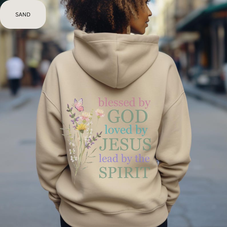 Pu&ograve; includere: Una felpa con cappuccio beige con un design floreale e il testo "blessed by GOD loved by JESUS lead by the SPIRIT" stampato sul retro.