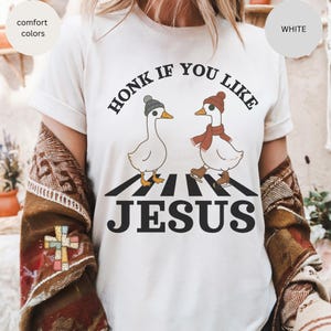 May include: White t-shirt with the text "HONK IF YOU LIKE JESUS". The graphic features two cartoon geese wearing hats and scarves, crossing a crosswalk. The shirt has comfort colors.