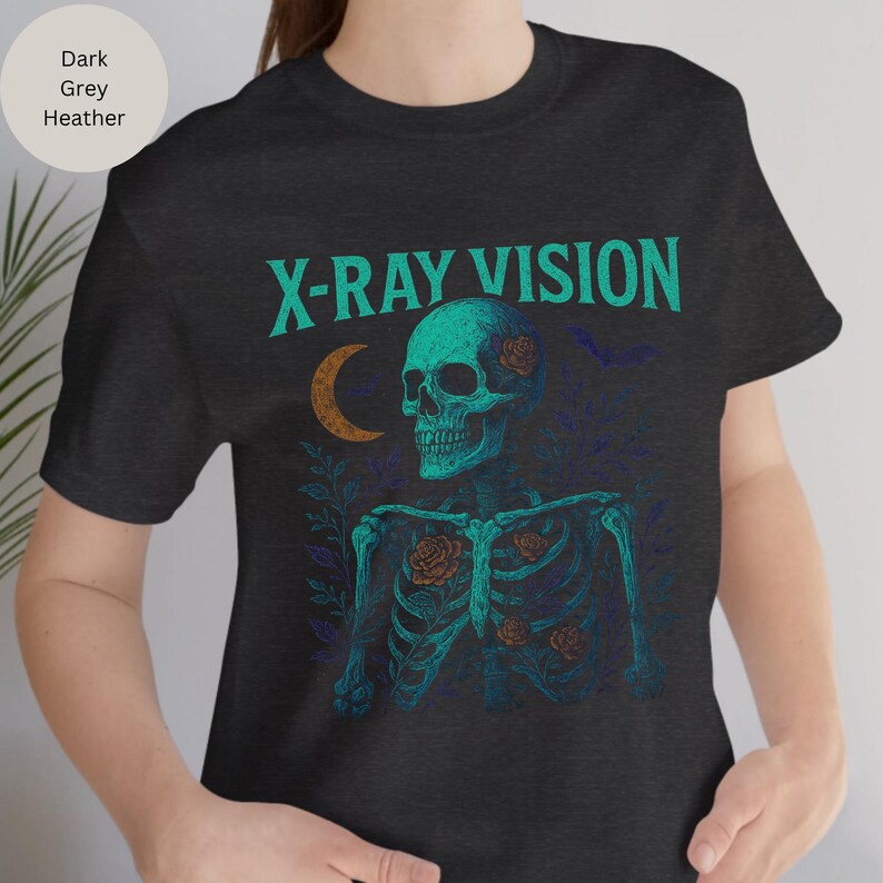 May include: Dark grey heather t-shirt with a teal and orange graphic. The graphic features a skeleton with roses, a crescent moon, and the words "X-RAY VISION" in teal. The design has a vintage, gothic aesthetic.