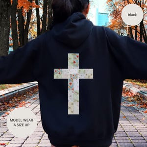 Puede incluir: Sudadera con capucha negra con un gran diseño de cruz en la espalda. La cruz está hecha de cuadrados de patchwork con telas florales y estampadas. También es visible el texto "MODEL WEAR A SIZE UP".