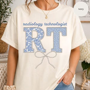 May include: Ivory-colored t-shirt with the words "radiology technologist" in blue. The letters "RT" are in a light blue floral pattern and dotted design, outlined with pearls. A pearl bow is below the letters.