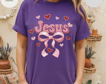 May include: A purple t-shirt with the word "Jesus" in pink, adorned with a pink bow and red hearts. The shirt has rolled-up sleeves and is labeled "comfort colors" and "Grape".