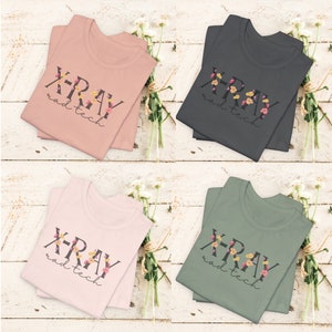 May include: Four folded t-shirts with the text "X-RAY rad tech" in black lettering with floral accents. The shirts are in four colors: pink, gray, light pink, and green.