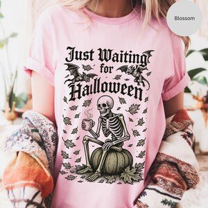 May include: Pink t-shirt with a Halloween-themed graphic. The design features a skeleton sitting on a pumpkin, holding a mug, with the text "Just Waiting for Halloween" and bat illustrations.