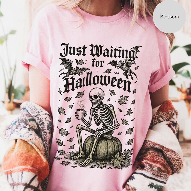 May include: Pink t-shirt with a Halloween-themed graphic. The design features a skeleton sitting on a pumpkin, holding a mug, with the text "Just Waiting for Halloween" and bat illustrations.