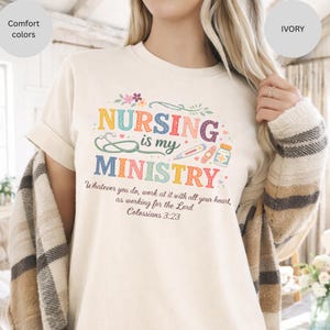 May include: Ivory-colored t-shirt with the text "NURSING is my MINISTRY" in colorful letters and medical-themed illustrations. The shirt also features the Bible verse Colossians 3:23.