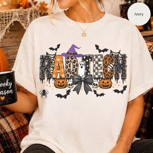 May include: An ivory t-shirt featuring the text RAD TECH in patterned letters, a purple witch hat, a white ghost, a black bow, two orange pumpkins, skulls, spiderwebs, spiders, and bats. The design incorporates leopard print, plaid, and checkered patterns.