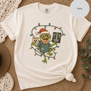 May include: An ivory short-sleeve t-shirt featuring a graphic of a green frog wearing a Santa hat, glasses, and a scarf. The frog holds a sign labeled RADIOLOGY and an X-ray image, surrounded by a string of colorful Christmas lights.