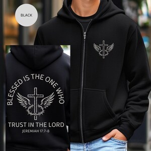 May include: Black zip-up hoodie with a white graphic of a cross, wings, and a crown of thorns. The back of the hoodie features the text "BLESSED IS THE ONE WHO TRUST IN THE LORD" and "JEREMIAH 17:7-8". The hoodie has a front pocket and a hood.