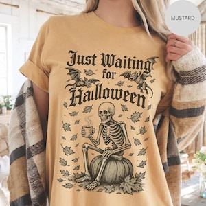 May include: Mustard-colored t-shirt with a Halloween-themed graphic. The design features a skeleton sitting on a pumpkin, holding a mug, with the text "Just Waiting for Halloween" and bats. The shirt has short sleeves and a relaxed fit.