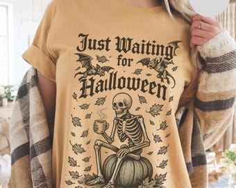 May include: Mustard-colored t-shirt with a Halloween-themed graphic. The design features a skeleton sitting on a pumpkin, holding a mug, with the text "Just Waiting for Halloween" and bats. The shirt has short sleeves and a relaxed fit.