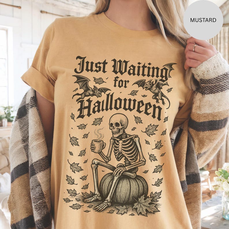 May include: Mustard-colored t-shirt with a Halloween-themed graphic. The design features a skeleton sitting on a pumpkin, holding a mug, with the text "Just Waiting for Halloween" and bats. The shirt has short sleeves and a relaxed fit.