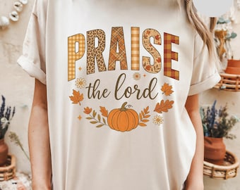 May include: Ivory-colored t-shirt featuring the words "PRAISE the lord" in a fall-themed design. The word "PRAISE" uses a mix of patterns, including plaid and leopard print. A pumpkin and fall leaves are also featured.