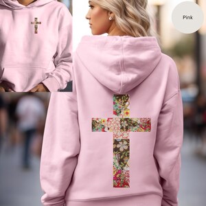 May include: A pale pink hoodie featuring a floral cross motif on the back and a smaller version on the front. The cross design incorporates a variety of colourful flowers set against a dark backdrop. The hoodie includes a hood and a front pocket.