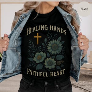 May include: Black t-shirt with the text "HEALING HANDS FAITHFUL HEART" in a gold font, featuring a gold cross and floral design in shades of green and blue. The shirt is a crew neck.