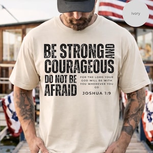 May include: Ivory-colored t-shirt with the text "BE STRONG AND COURAGEOUS DO NOT BE AFRAID" in bold black letters. The text also includes the Bible verse Joshua 1:9. The shirt is a casual wear item.
