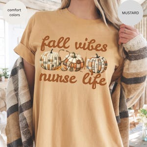 May include: Mustard-colored t-shirt with the text "fall vibes nurse life" in brown script. The design features three pumpkins with patchwork patterns and a stethoscope. The shirt is a comfort color.