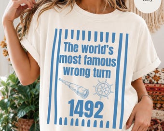Könnte beinhalten: Ein elfenbeinfarbenes T-Shirt mit einem blauen Grafikdesign mit vertikalen Streifen. Der Text lautet "The world's most famous wrong turn 1492", ergänzt durch Illustrationen eines nautischen Fernrohrs und eines Schiffssteuerrads in Blau.