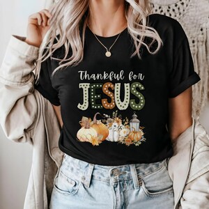 Op de afbeelding: Zwart T-shirt met de tekst "Thankful for JESUS" in kleurrijke letters met stippen. De tekst wordt omringd door herfstthema-pompoenen, bloemen en een lantaarn.
