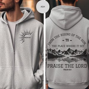 May include: Ash grey zip-up hoodie. The front has a black sunburst graphic and the text "PSALM 113:3". The back features a mountain and lake scene with the text "FROM THE RISING OF THE SUN TO THE PLACE WHERE IT SET PRAISE THE LORD PSALM 113:3".