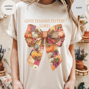 May include: Ivory-colored t-shirt with the text "GIVE THANKS TO THE LORD" above a large bow illustration. The bow features a patchwork design with pumpkins, grapes, and floral patterns in shades of orange, pink, and purple.