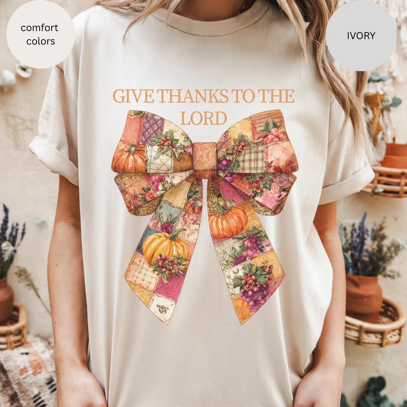 May include: Ivory-colored t-shirt with the text "GIVE THANKS TO THE LORD" above a large bow illustration. The bow features a patchwork design with pumpkins, grapes, and floral patterns in shades of orange, pink, and purple.