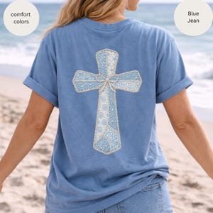 May include: A blue t-shirt with a cross design on the back. The cross is light blue and white, with a bow at the center. The shirt has rolled-up sleeves and the words "comfort colors" and "Blue Jean" are in the image.
