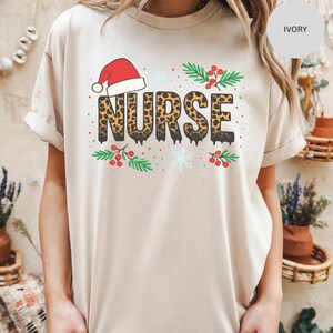 May include: Ivory-colored t-shirt with the word "NURSE" in leopard print, topped with a Santa hat. The design includes green sprigs with red berries and snowflakes, creating a festive holiday theme.