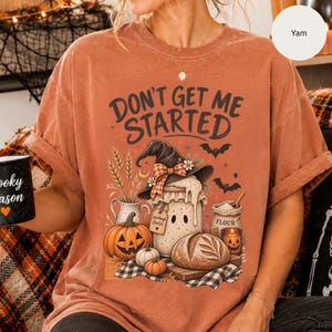 May include: A rust-colored short-sleeve t-shirt featuring a graphic print with the text "DON'T GET ME STARTED" above a witch hat, a jar labeled "sourdough starter," pumpkins, a loaf of bread, a bag of flour, and a candle, all set on a checkered cloth.