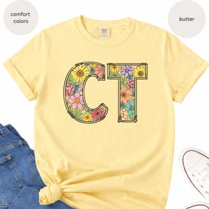 May include: A light yellow t-shirt with the letters "CT" filled with colorful floral designs. The shirt is made of a soft material and has short sleeves. The shirt is shown with blue jeans and white sneakers.