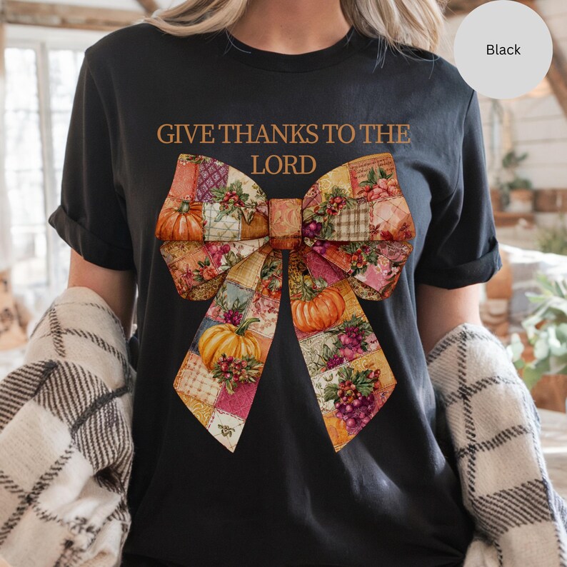 May include: Black t-shirt with the text "GIVE THANKS TO THE LORD" in a brown font. The shirt features a large bow design with a patchwork pattern, pumpkins, and grapes in shades of orange, yellow, and purple.