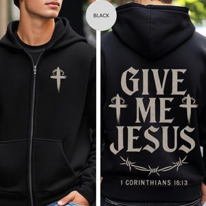 May include: Black zip-up hoodie. The front has a silver cross design, while the back displays "GIVE ME JESUS", a barbed wire design, and the Bible verse 1 Corinthians 16:13.