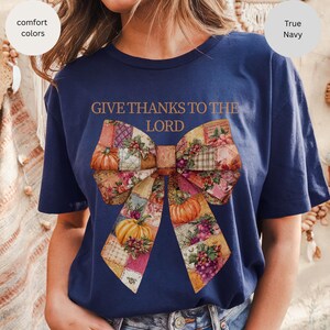 May include: Navy blue t-shirt with the text "GIVE THANKS TO THE LORD" above a large bow design. The bow features a patchwork pattern with pumpkins, flowers, and grapes. The shirt has short sleeves and is made of a soft material.