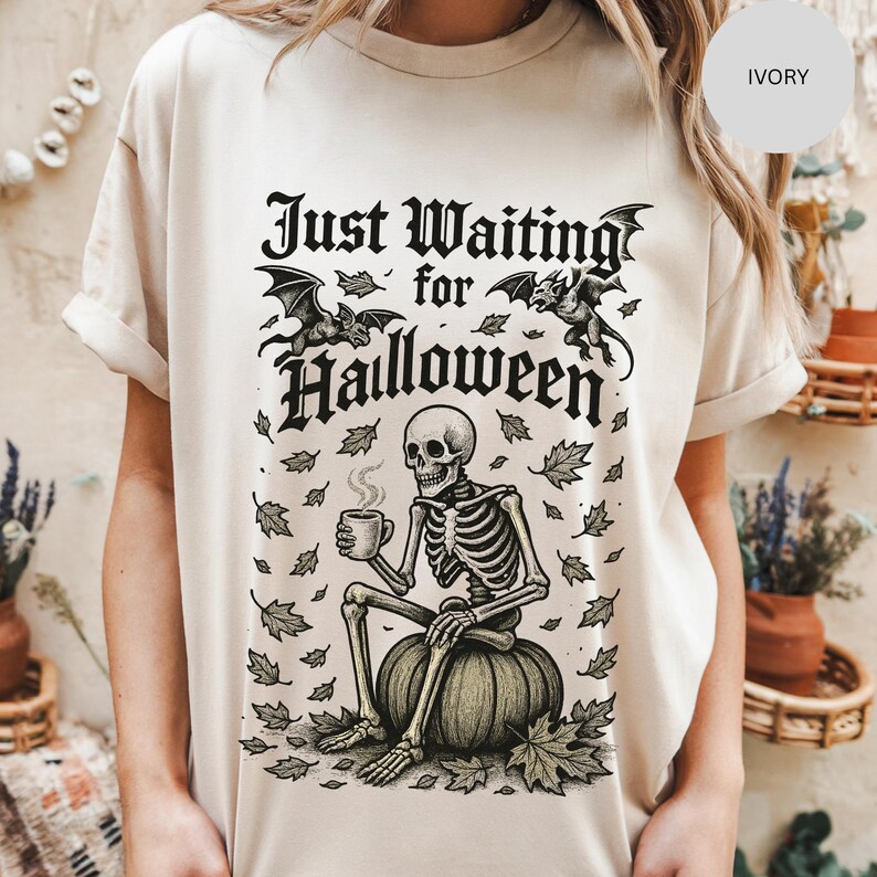 May include: Ivory-colored t-shirt with a Halloween-themed graphic. The design features a skeleton sitting on a pumpkin, holding a mug, with the text "Just Waiting for Halloween" above. Bats and leaves are also included in the design.