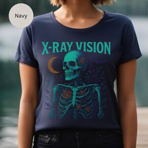 May include: Navy blue t-shirt featuring a teal skeleton with roses and the text "X-RAY VISION" in teal. The design includes a crescent moon and floral elements. The shirt is a crew neck.