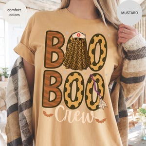 May include: Mustard-colored t-shirt with a Halloween-themed design. The design features the words "BOO BOO Crew" in a mix of fonts and patterns, including leopard print and zebra stripes. A witch's hat and broom are also included in the design.
