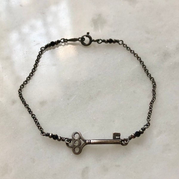 May include: A silver key charm bracelet with a delicate chain and black beads. The key charm has an ornate design and a small letter 'F' on the end.