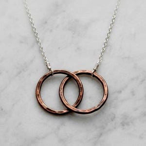 May include: A silver chain necklace featuring two interlocking copper-colored rings. The rings have a hammered texture and are connected to the chain with silver wire. The necklace is displayed on a white marble surface.