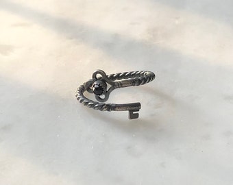 May include: A silver ring with a twisted band and a key charm with a black gemstone. The key charm is in the shape of an old-fashioned key with a round head.
