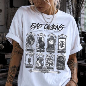 May include: A white short-sleeve t-shirt featuring a black graphic print titled A Field Guide To Bad Omens. The design includes eight rectangular illustrations depicting a broken mirror, a black cat, a clock, a beetle, a candle, a mailbox, flowers, and an umbrella.