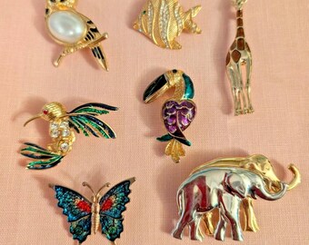 May include: A selection of vintage animal brooches. The collection features an owl, fish, giraffe, hummingbird, toucan, butterfly, and elephant. The brooches are gold, silver, and jewel-toned, set against a pink background.