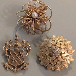 Può includere: Tre spille color oro: una presenta una perla centrale circondata da anelli di filo radianti, un'altra è uno stemma a forma di scudo con volute ornamentali e la terza è un pezzo testurizzato a forma di fiore con piccole pietre chiare.