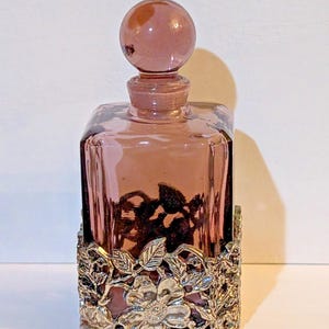 Puede incluir: Un frasco de perfume vintage de vidrio malva, de forma cuadrada, con un tapón redondo. Se encuentra en una base de filigrana de metal plateado adornada con motivos florales y de hojas. La botella mide aproximadamente 15 cm de altura.