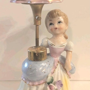 May include: A ceramic figurine of a person wearing a pink, blue, and yellow dress with gold accents. The figure holds a metal pole topped with a pink umbrella-shaped piece. The dress features small painted floral details and gold trim.