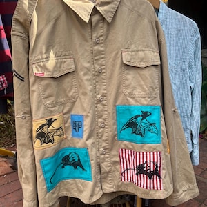 May include: A tan long-sleeve button-down shirt featuring two chest pockets, shoulder epaulets, and four rectangular patches with black ink illustrations of bats and an opossum. A small blue patch reads HEY TAYLOR CAGE.